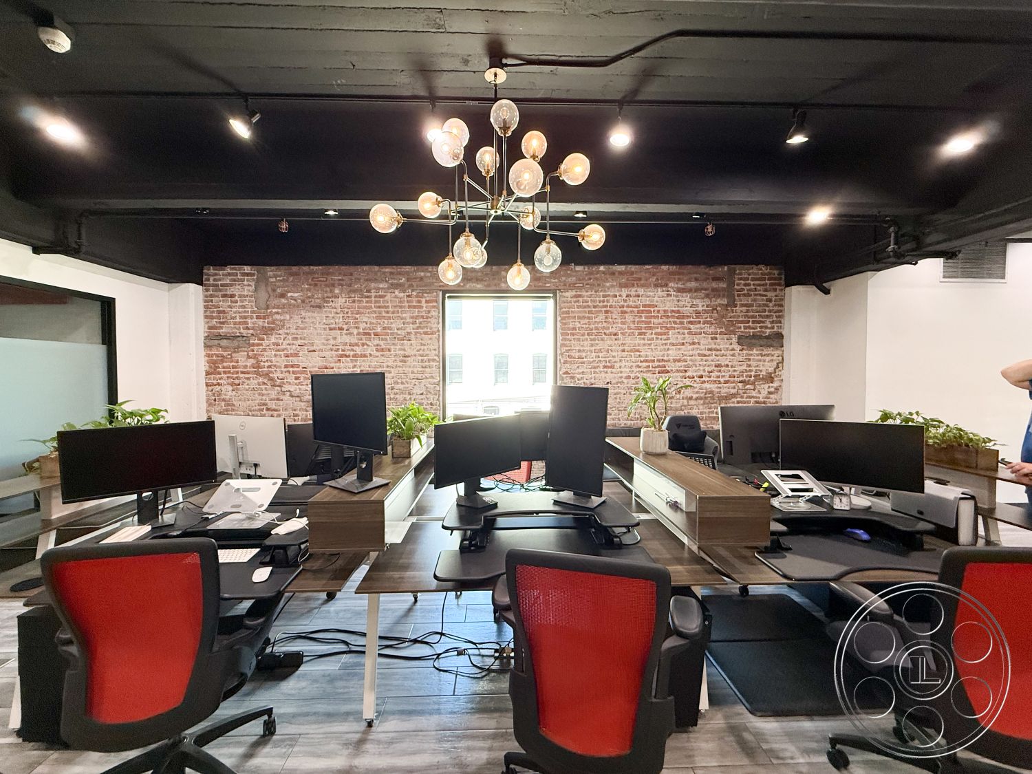Office 124 - exposed brick accent wall interior, black painted ceiling interior, wood desk with integrated storage interior, luminaire chandelier with glass globes interior, computer workstation setup interior, red ergonomic office chairs interior, large windows with city view interior, potted greenery accents interior, laminate flooring with wood texture interior, modern office design interior, industrial style office interior, open-plan workspace interior, minimalistic office decor interior, transparent glass partitions interior