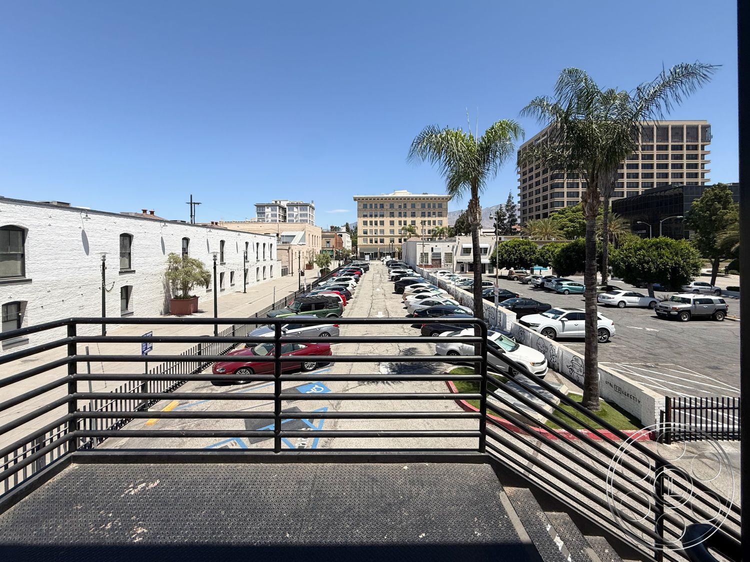Office 124 - clear blue sky, industrial-style architecture, exterior concrete parking lot, exterior asphalt roadway, exposed brick warehouse walls, metal railings on elevated walkway, lush palm trees in urban setting, mid-century modern building aesthetics, urban skyline view in background, hardscape design with concrete planters, straight parking layout, suburban city environment, urban commercial district, decorative light fixtures on street, mixed-use development area, historic building facades, pedestrian walkway with metal grates