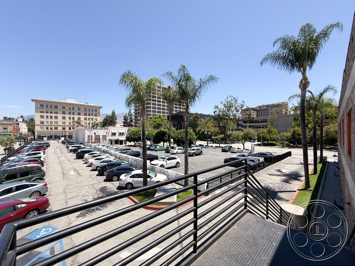 Office 124 - exterior view, urban setting, open-air environment, mixed-use development, landscaped green areas, palm tree landscaping, concrete parking surface, city skyline backdrop, metal stair railings, urban parking lot, two-story commercial building, brick building facade, flat roof architecture, asphalt road surface, sidewalk with ADA accessible parking, modern multi-story buildings, bright sunny day setting