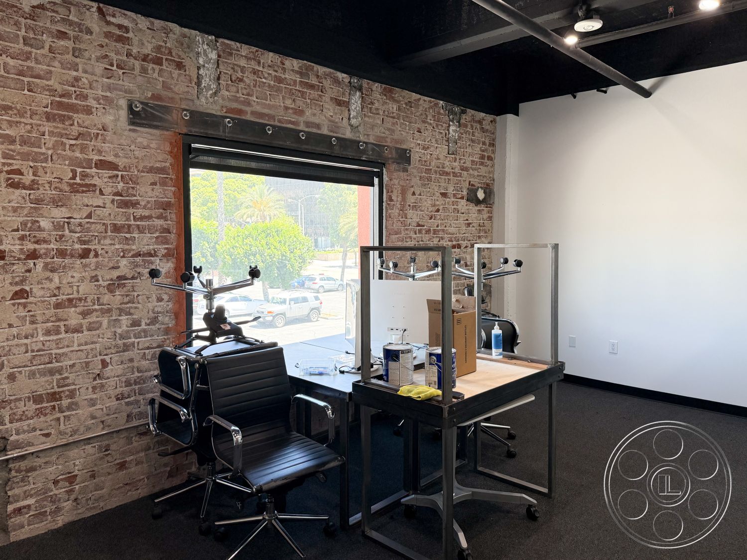Office 124 - floor-to-ceiling windows interior, exposed brick accent wall interior, natural light from large windows interior, black steel structural beams interior, open workspace layout interior, black office chairs interior, padded office chairs interior, glass desk tops interior, rolling cart workspace interior, dark carpet flooring interior, sleek industrial design interior, minimalist office decor interior, urban loft workspace setting, city view from windows interior