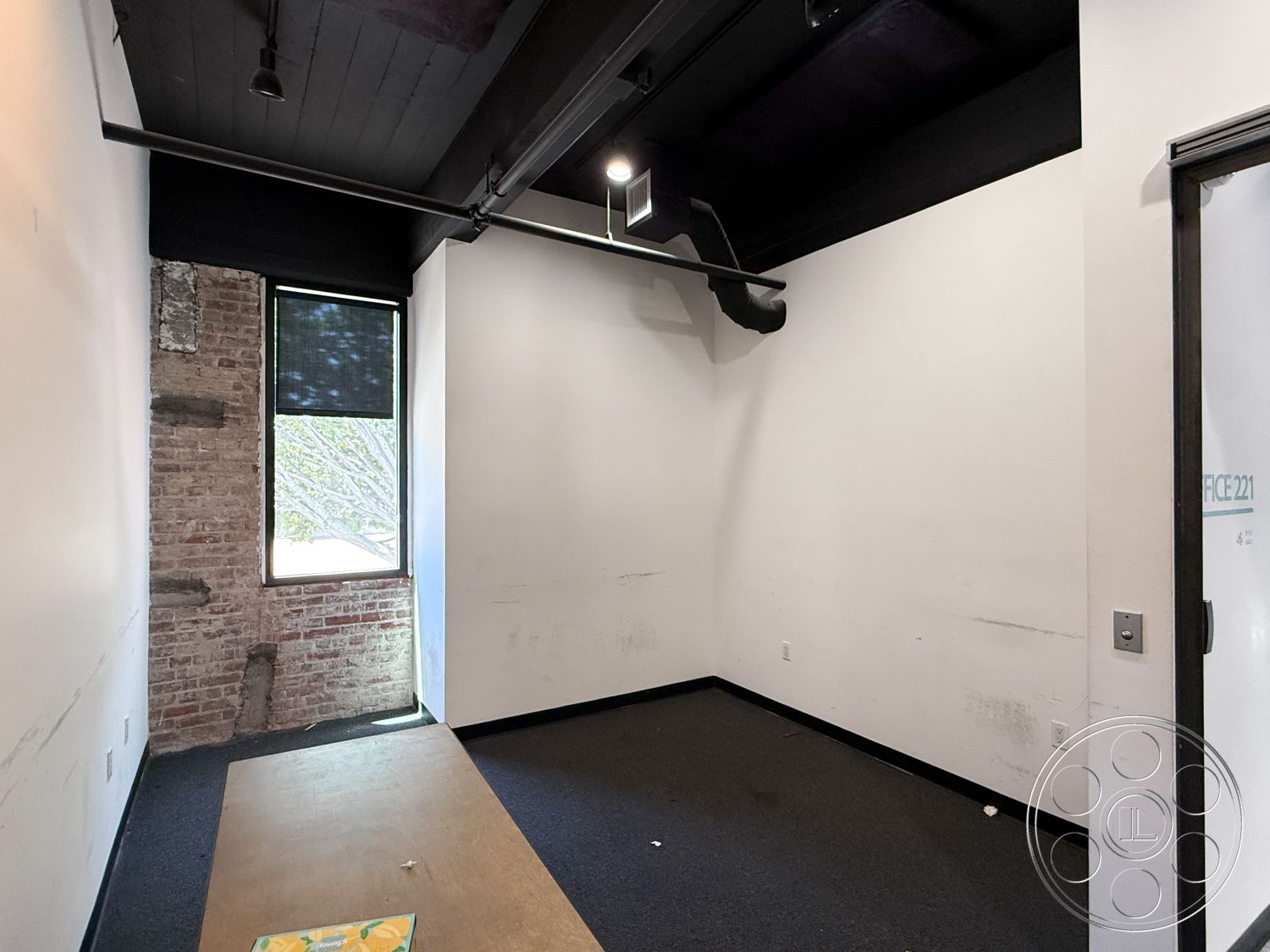 Office 124 - exposed brick accent wall interior, industrial style interior, black ceiling beams interior, flat black ceiling interior, black drywall interior, carpet flooring interior, large window with exterior view, minimalist office space interior, modern office layout interior, urban office setting interior