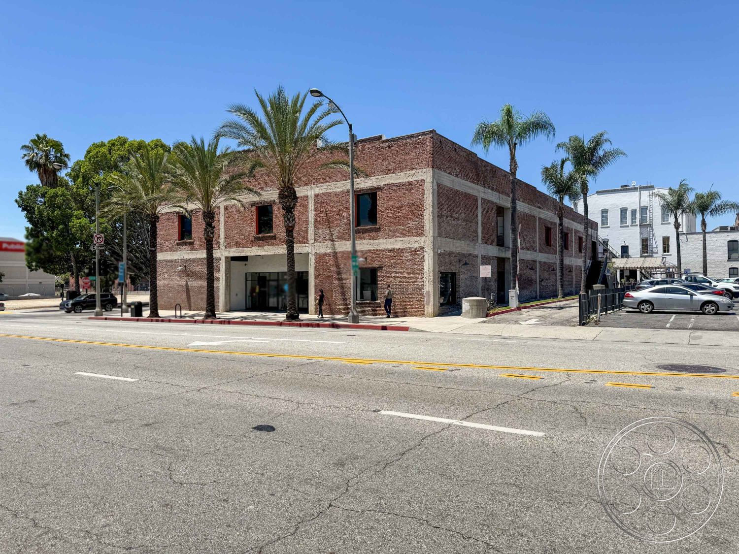 Office 124 - modern entrance, surrounding palm trees, two-story structure, concrete sidewalk, flat roof design, palm tree landscaping, city street setting, industrial style architecture, brick exterior walls, painted concrete foundation, large glass storefront windows, urban commercial building, exposed brick accent, street-facing parking lot, downtown area