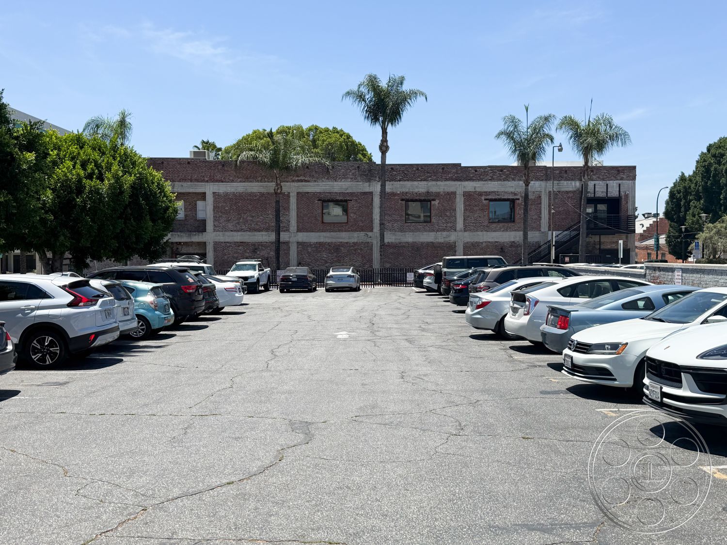 Office 124 - urban setting, clear blue sky, brick building facade, exterior parking lot, asphalt parking surface, landscape with palm trees, two-story industrial building, fenced parking area, metal fire escape, diverse parked vehicles