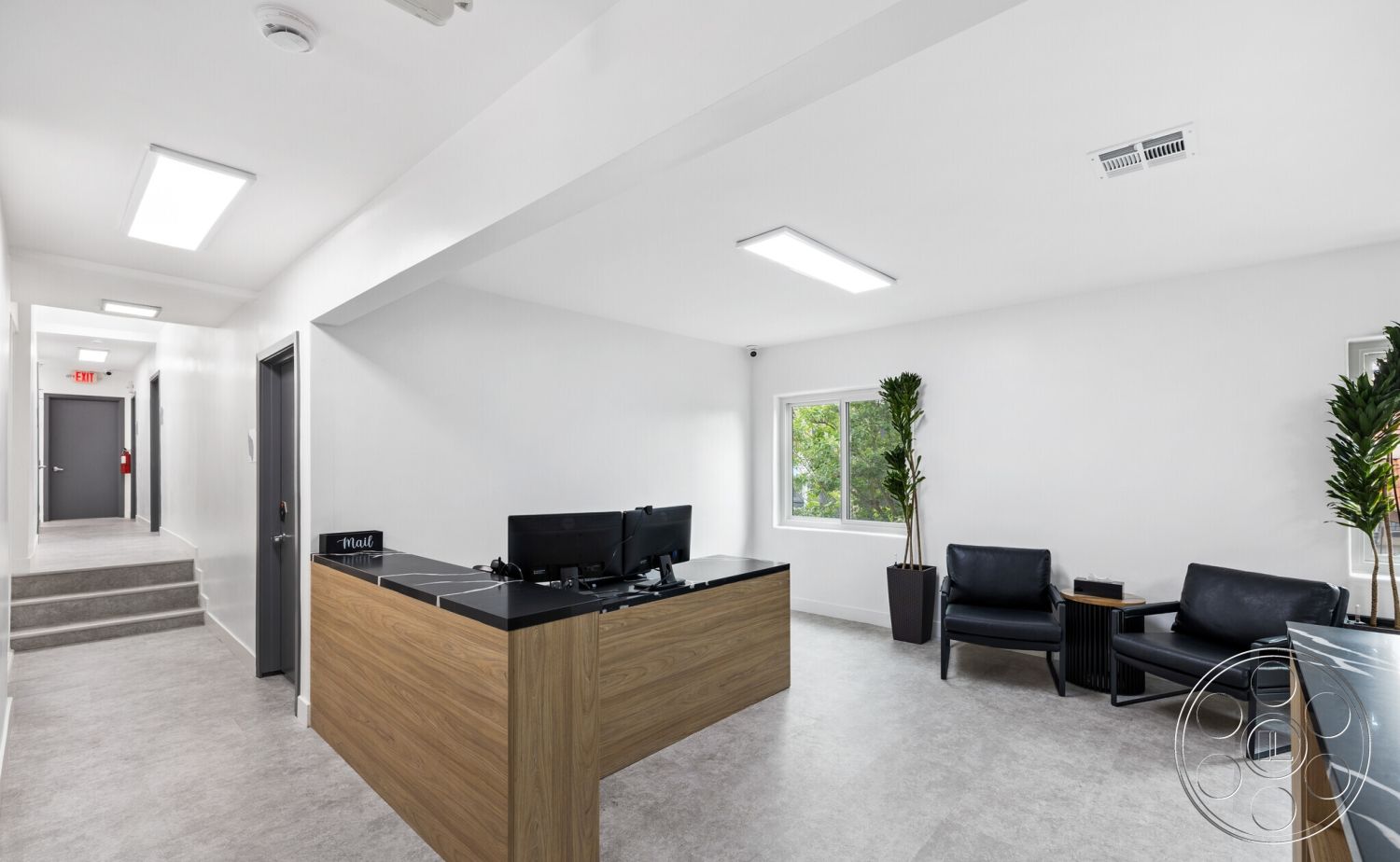 Office 125 - neutral color palette, interior space, decorative potted plants, white painted walls, bright and airy ambiance, minimalist architectural style, track lighting fixtures, smooth surface finishes, textured ceiling panels, light gray vinyl flooring, wood reception desk, black countertop on reception desk, large window with modern trim, black leather seating, metal door hardware, contemporary artwork on walls, clean lines and open layout, step up hallway to additional rooms, functional office environment
