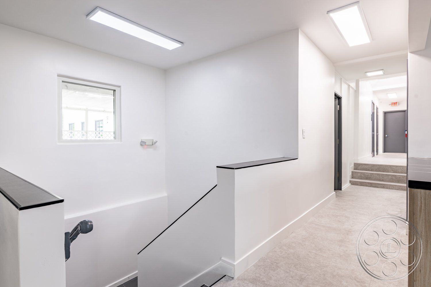 Office 125 - open layout, modern lighting fixtures, minimalist design elements, interior hallway, white painted walls, smooth wall texture, staircase with metal railings, concrete floor tiles, gray carpeted stairs, small window with security features, functional circulation space