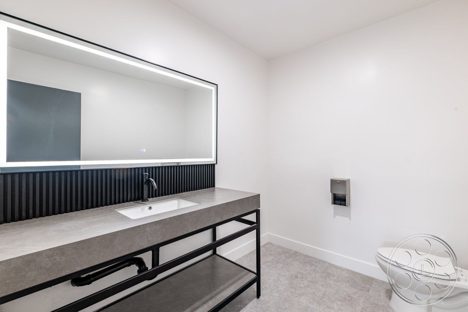 Office 125 - bathroom, open space layout, backlit mirror, white painted walls, modern minimalist design, interior bathroom, concrete countertop sink, black metal bathroom vanity, textured black wall paneling, light gray porcelain floor tiles, contemporary faucet fixtures, flush mount ceiling lighting, simple toilet design, stylish toilet paper holder
