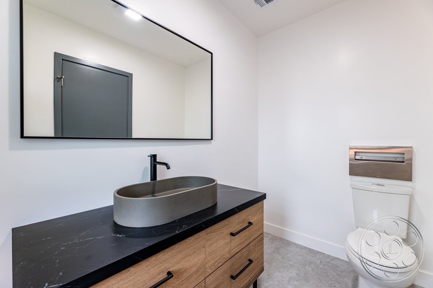 Office 125 - bathroom, minimalist design, black marble countertop, white painted walls, interior bathroom, modern faucet fixtures, oval stone sink, wood grain vanity cabinets, dark grey door, large rectangular mirror, contemporary wall lighting, light grey concrete flooring, modern bathroom aesthetics