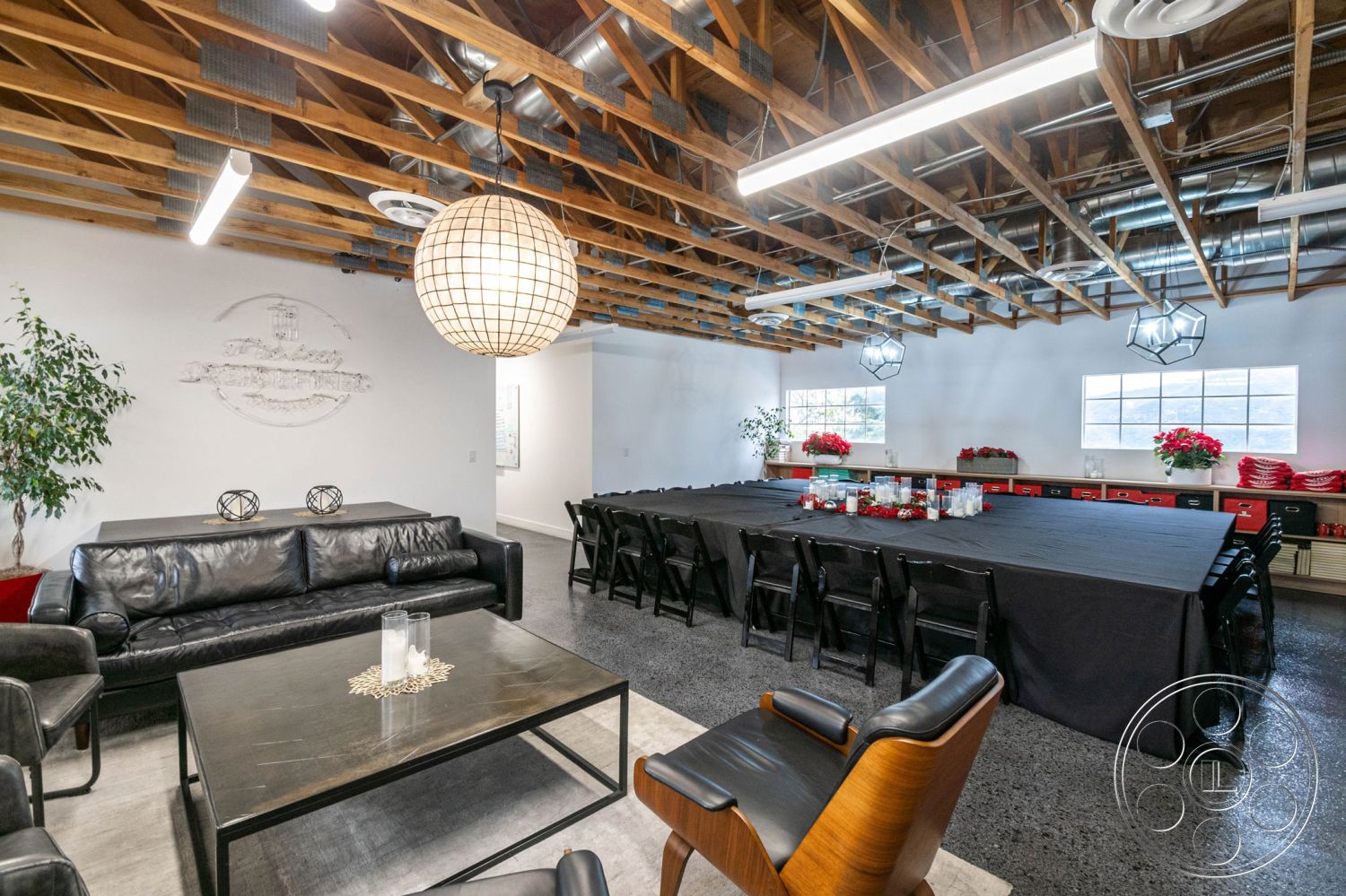 Office 126 - concrete flooring, modern interior, urban setting, warm ambient lighting, exposed wood ceiling beams, large windows with garden view, white painted walls, functional meeting space, linear pendant lighting fixtures, metal ductwork ceiling details, black leather sectional sofa, glass coffee table with gold accents, long rectangular dining table, black folding chairs, decorative potted plant