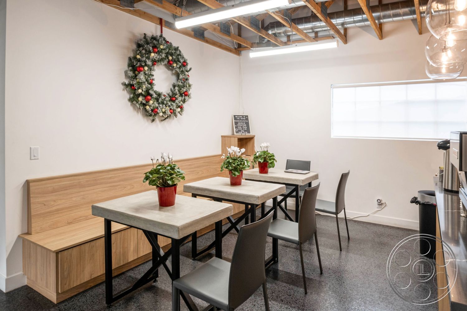 Office 126 - polished concrete flooring, open concept layout, exposed wood ceiling beams, interior dining area, minimalist modern decor, white painted walls, industrial-style lighting fixtures, decorative holiday wreath, concrete dining table, black steel table legs, light wood bench seating, spacious natural light window, potted green plants, designer shelving unit