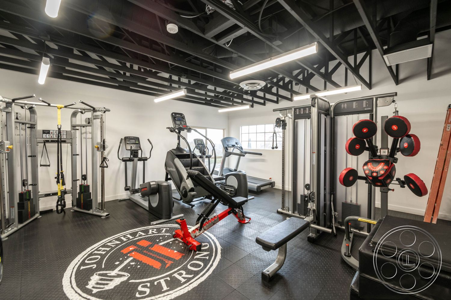 Office 126 - large windows for natural light, black rubber flooring, cardio machines, white painted walls, industrial-style decor, exposed black ceiling beams, interior gym space, rubber floor tiles, fitness equipment area, strength training equipment, LED strip lighting, functional home gym