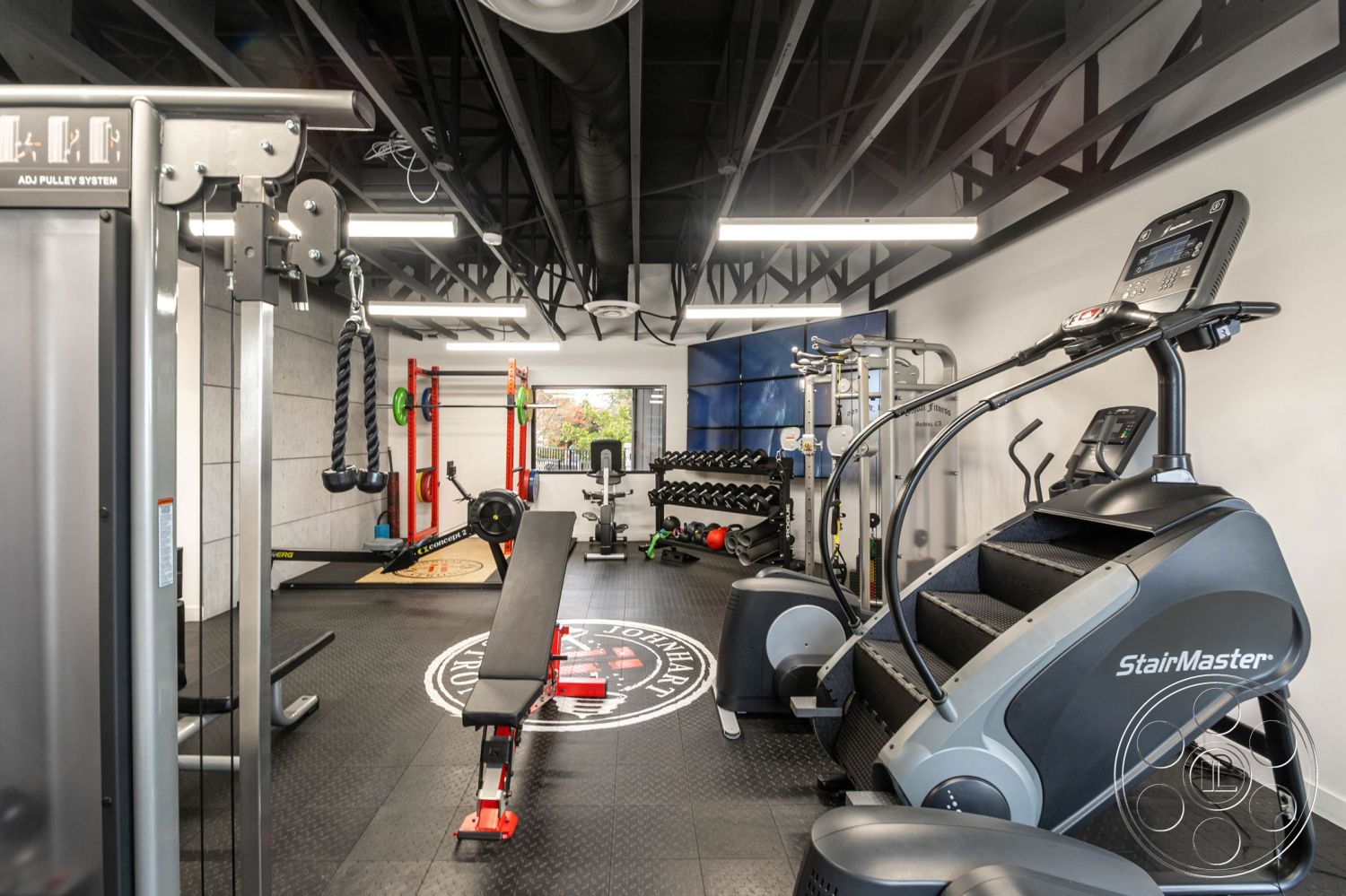 Office 126 - cable machine, cardio machines, large windows with natural light, exposed metal ceiling beams, rubber gym flooring, interior gym space, concrete block accent wall, industrial-style gym equipment, black rubber weightlifting mats, steel weight racks, LED overhead lighting, flat workout benches, adjustable pulley system, multifunctional gym station, wall-mounted plate storage, fitness studio with wall mirrors, open layout gym, functional training area, flat-screen display for workouts, modern aesthetic gym design