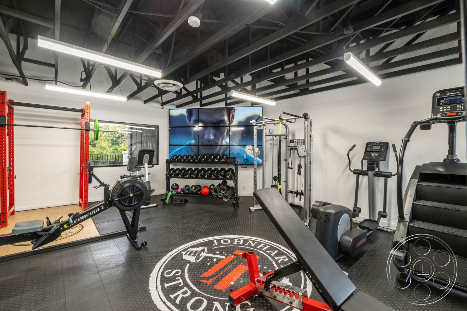 Office 126 - white painted walls, exposed metal ceiling beams, rubber gym flooring, adjustable weight bench, strength training equipment, black rubber tiles gym floor, open concept fitness room interior, fitness equipment storage rack, large wall-mounted television, cardio workout area with treadmill, bright LED strip lighting, large window with outdoor view, modern industrial gym decor, functional fitness space, residential home gym interior, high-tech workout station