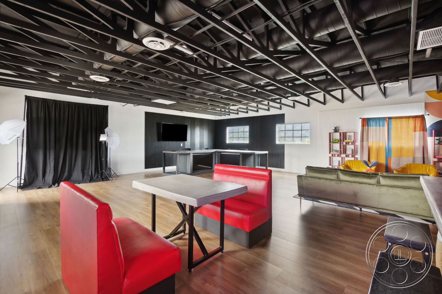 Office 126 - open floor plan, minimalist design elements, interior space, floor-to-ceiling windows, soft lighting fixtures, modern seating area, walnut hardwood flooring, contemporary dining table, industrial style decor, exposed black metal ceiling beams, red vinyl banquette seating, black metal table base, textured black accent wall, colorful fabric curtains, light wood bar counter, multicolored decorative shelving, studio setup with photography backdrop