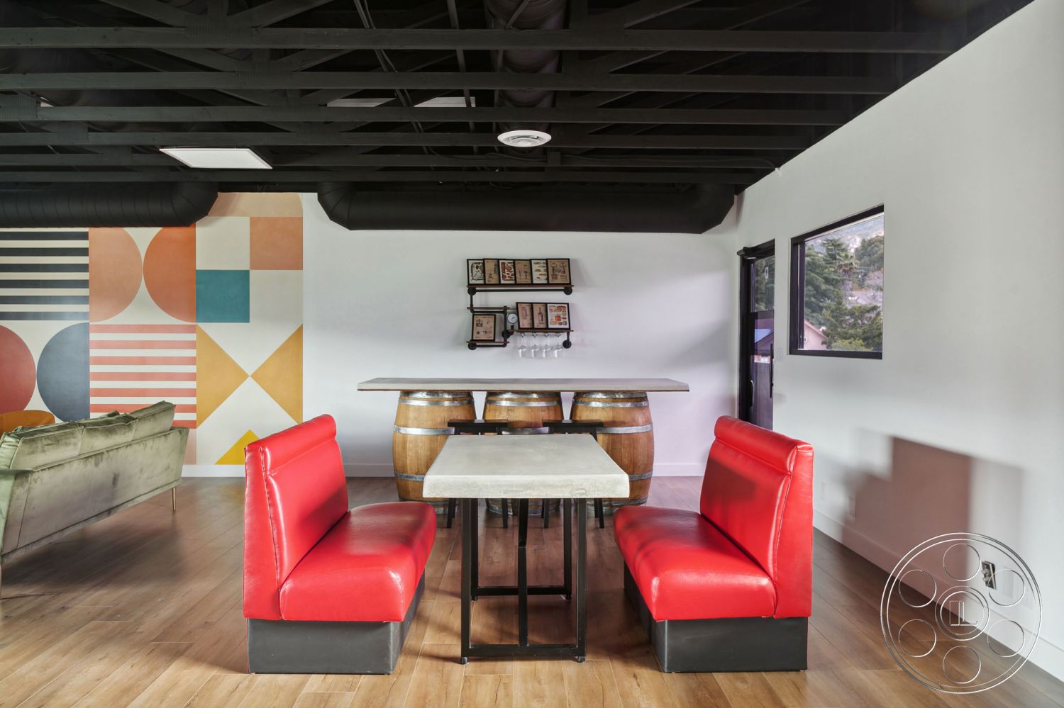 Office 126 - light hardwood flooring, colorful accent wall, contemporary seating area, open floor plan living area, modern industrial style, large windows with black frames, red vinyl booth seating, black exposed beam ceiling, concrete tabletop, wooden barrel tables, textured plaster wall with geometric mural, black metal shelving unit, functional dining space
