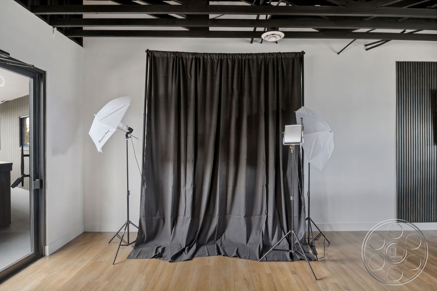 Office 126 - neutral color palette, textured wall paneling, open concept space, modern minimalist design, interior hardwood flooring, white painted walls, black curtain backdrop, studio lighting equipment, functional workspace environment, exposed black metal ceiling beams, large glass door entry, photography studio setup, natural light source from doorway, clean lines architectural details, versatile creative space