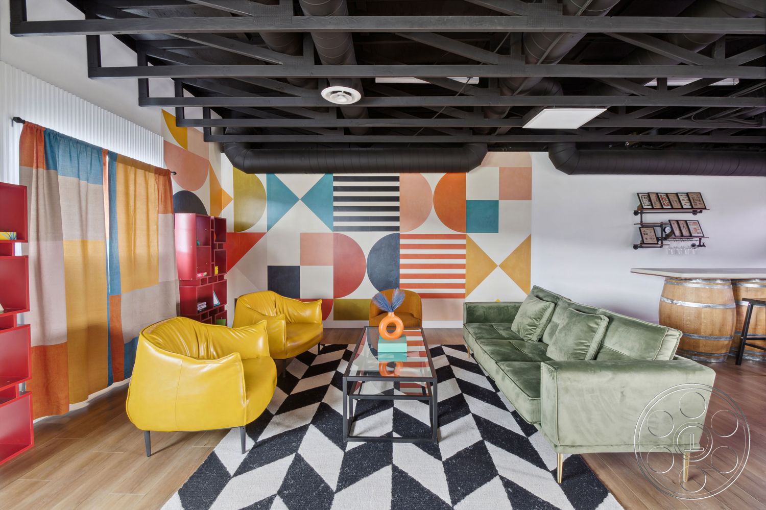 Office 126 - open layout, creative workspace, modern coffee table, contemporary design elements, green velvet sofa, yellow accent chairs, interior living room, wooden floorboards, exposed black metal ceiling beams, geometric mural wall art, colorful patterned curtains, black and white checkered area rug, red modular bookshelves, industrial-style bar area, multicolored decor accents