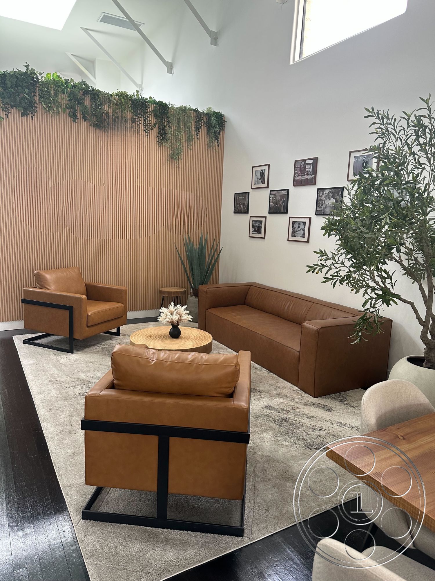 Office 127 - neutral color palette, natural light, area rug, leather armchair, textured wall paneling, hardwood floor, circular coffee table, leather sofa, framed black and white photographs, lush indoor plants, interior seating area, decorative indoor tree, metal base furniture, vertical slat wood accent wall, modern design style, open ceiling structure