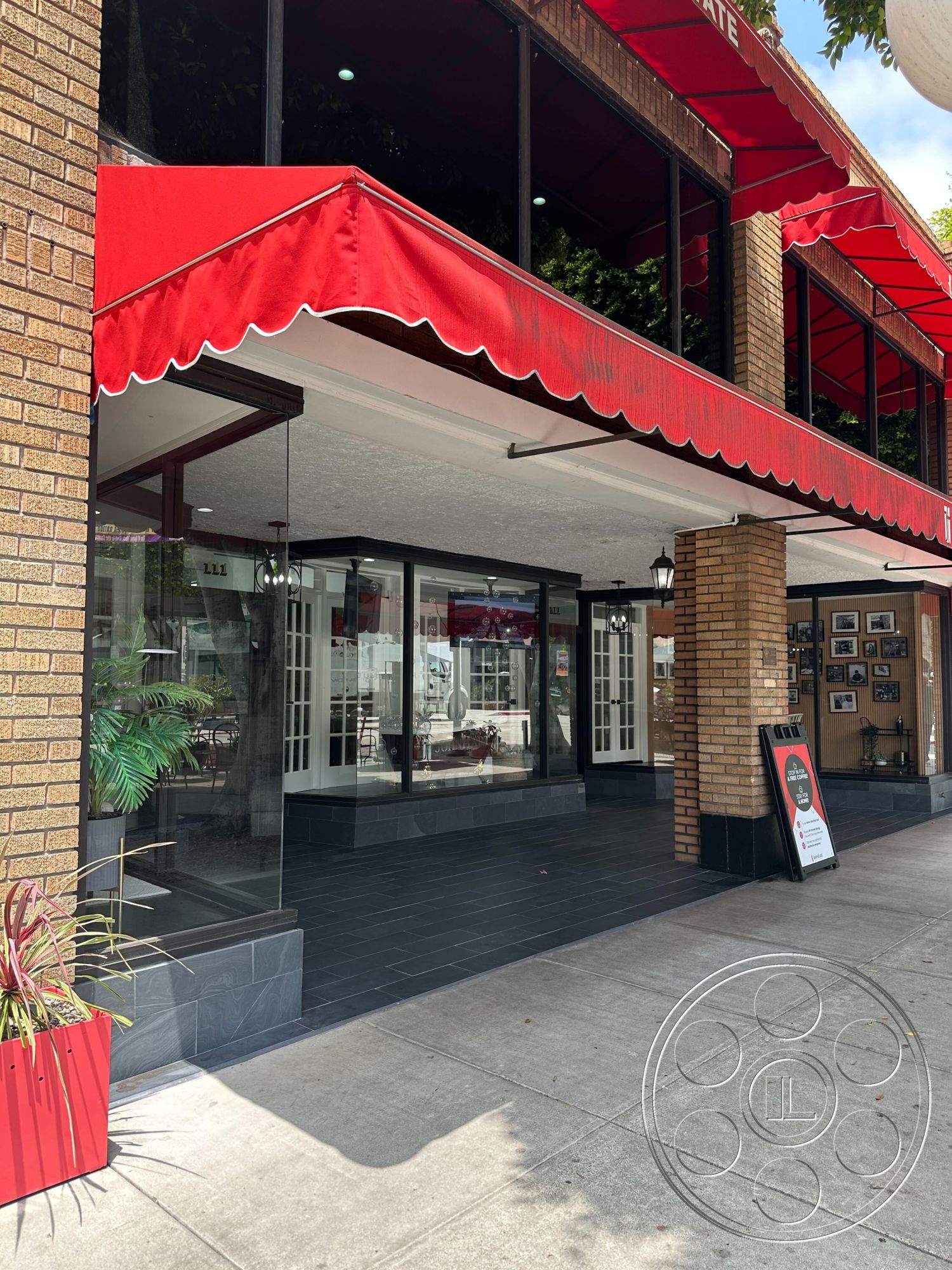Office 127 - black tile flooring, contemporary architectural design, glass storefront windows, city environment, exterior brick facade, red awning over storefront, exposed brick columns, planter with tropical plants, pedestrian sidewalk setting, business entrance with double doors, outdoor signage board, modern urban storefront, lamps hanging from ceiling, natural light streaming in