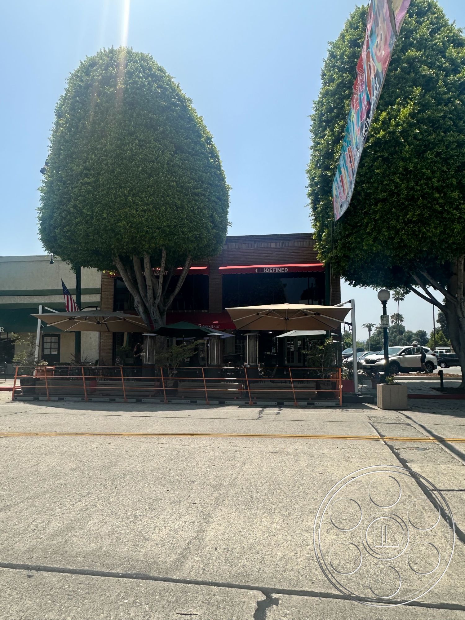 Office 127 - exterior, outdoor seating area, commercial building, decorative planters, red accent lighting, concrete sidewalk, pedestrian-friendly environment, brick building facade, large round canopy trees, rectangular patio space, metal railing fence, sheltered dining area, urban street setting, centralized location, restaurant signage, large awning structures