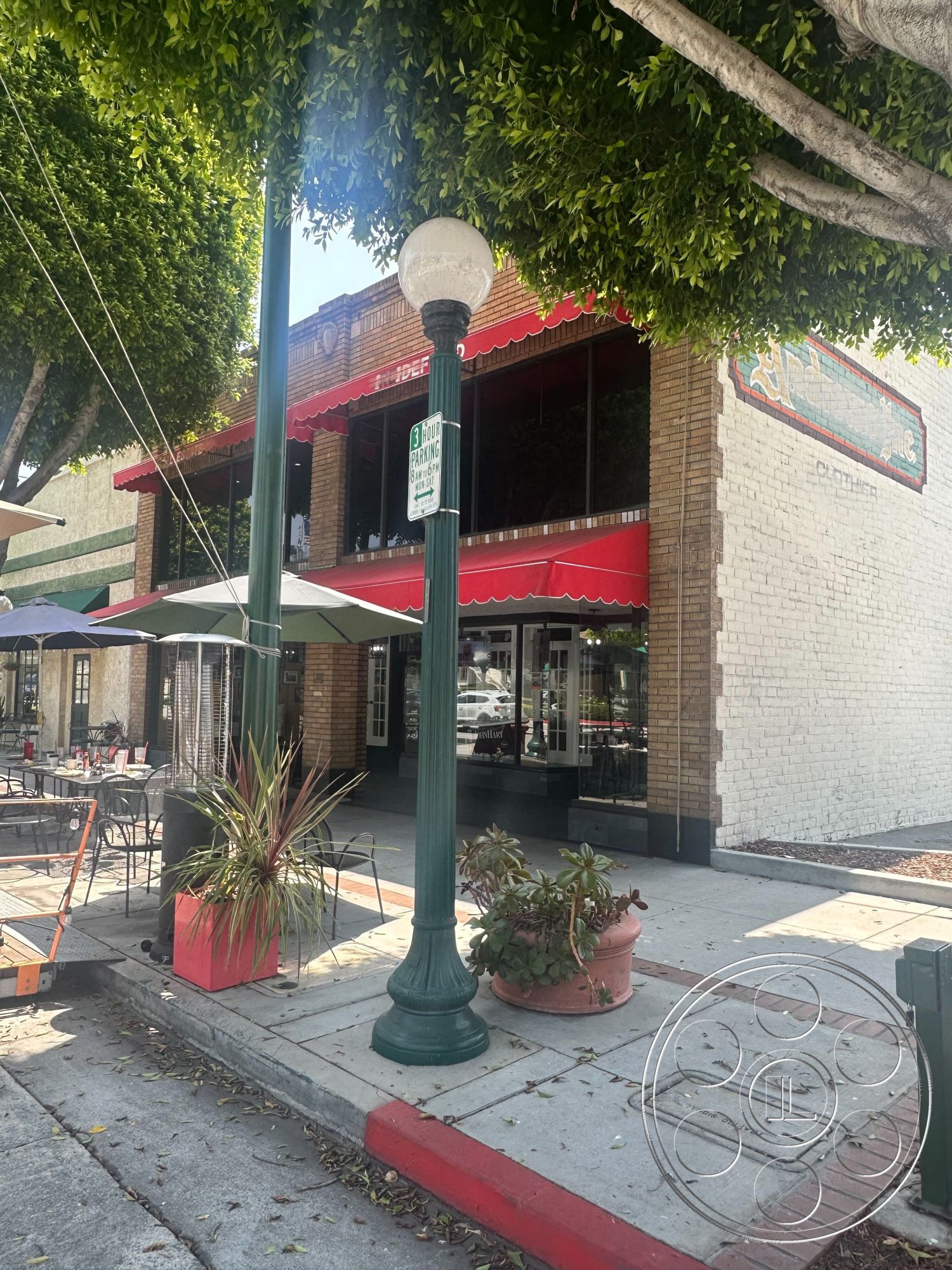 Office 127 - decorative lamppost, brick facade building exterior, green awning outdoor dining area, textured plaster wall accents, hardscape sidewalk design, red painted curbing, outdoor seating patio, potted ornamental plants, modern bistro table and chairs, urban streetscape setting, historic building architecture, brick column details, curved sidewalk edges, street-side parking designation sign, lush tree canopy overhead, textured tree bark, urban parklet atmosphere