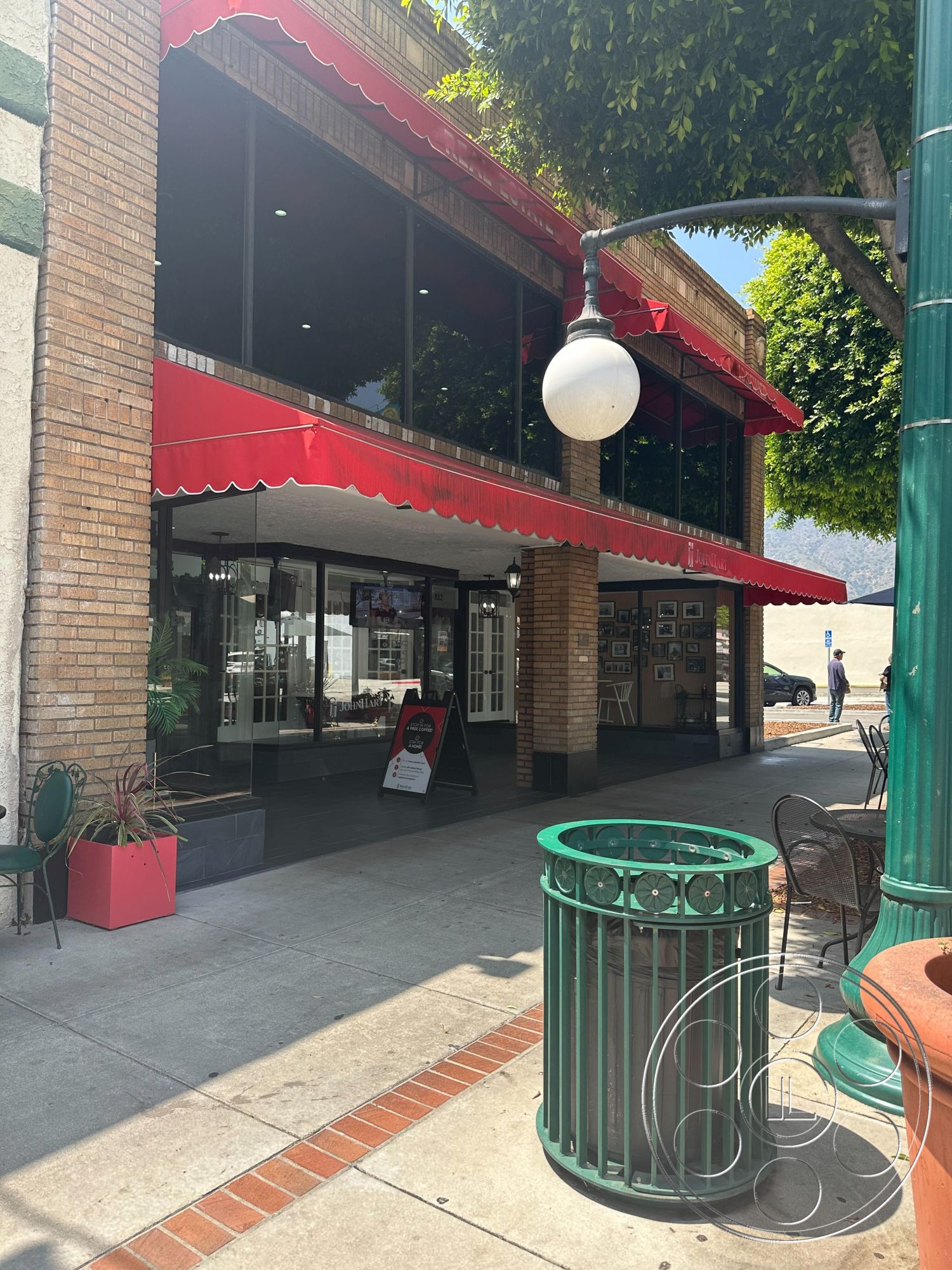 Office 127 - outdoor seating area, traditional architectural style, commercial building entrance, glass storefront windows, brick exterior walls, urban street setting, red awning, paved concrete walkway, decorative green metal trash can, circular sidewalk pattern, exposed brick architecture, landscaped exterior planters, sectioned opening design, pedestrian-friendly streetscape, brightly lit outdoor space, historical building facade