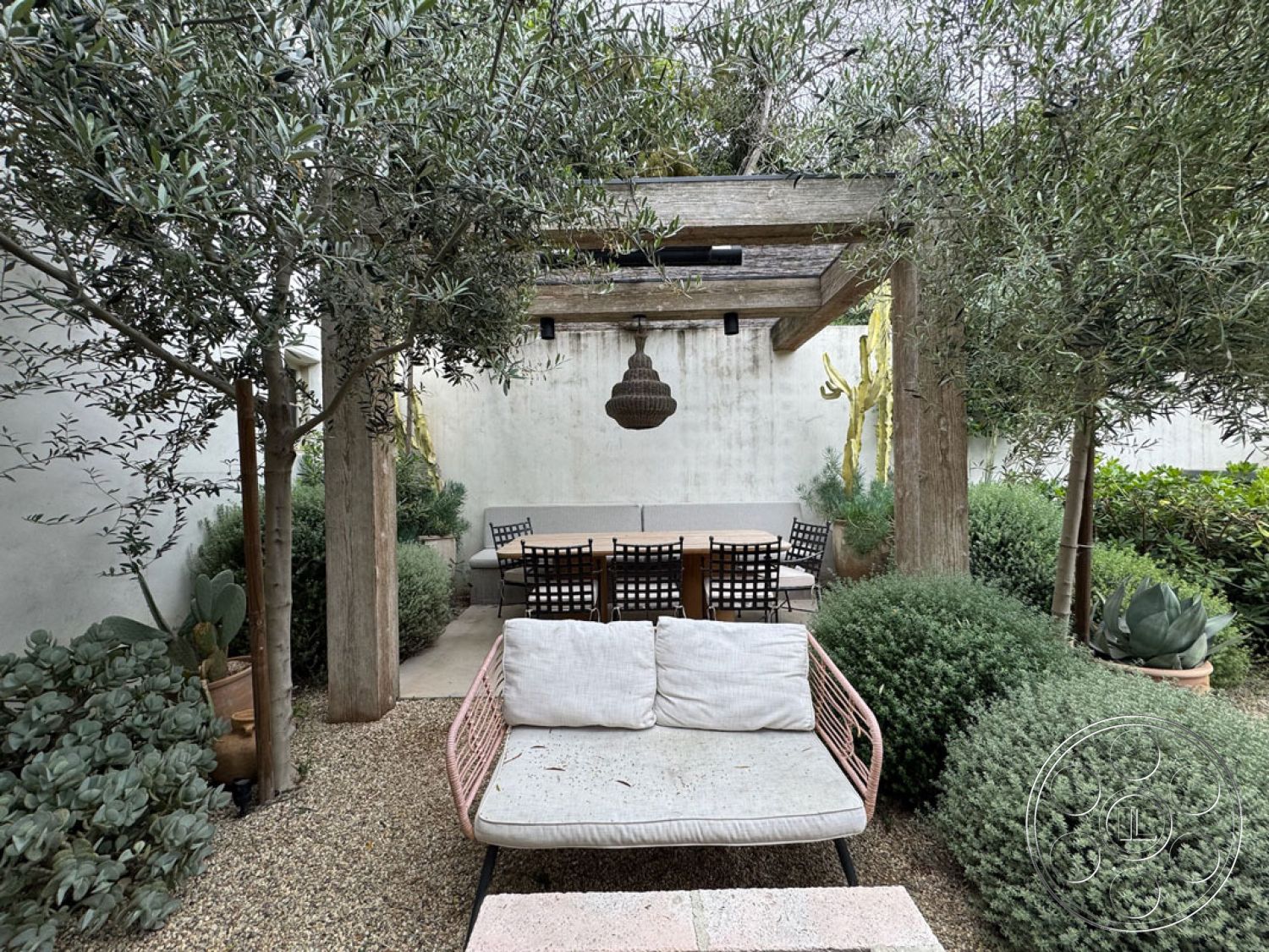 Spanish 165 - rustic outdoor dining area, concrete patio flooring, wood pergola structure, black metal dining chairs, exterior garden patio, lush greenery landscaping, modern outdoor lounge sofa, decorative pendant light fixture, potted succulent plants, textured white plaster wall, gravel garden floor, privacy wall with foliage, natural wood support beams