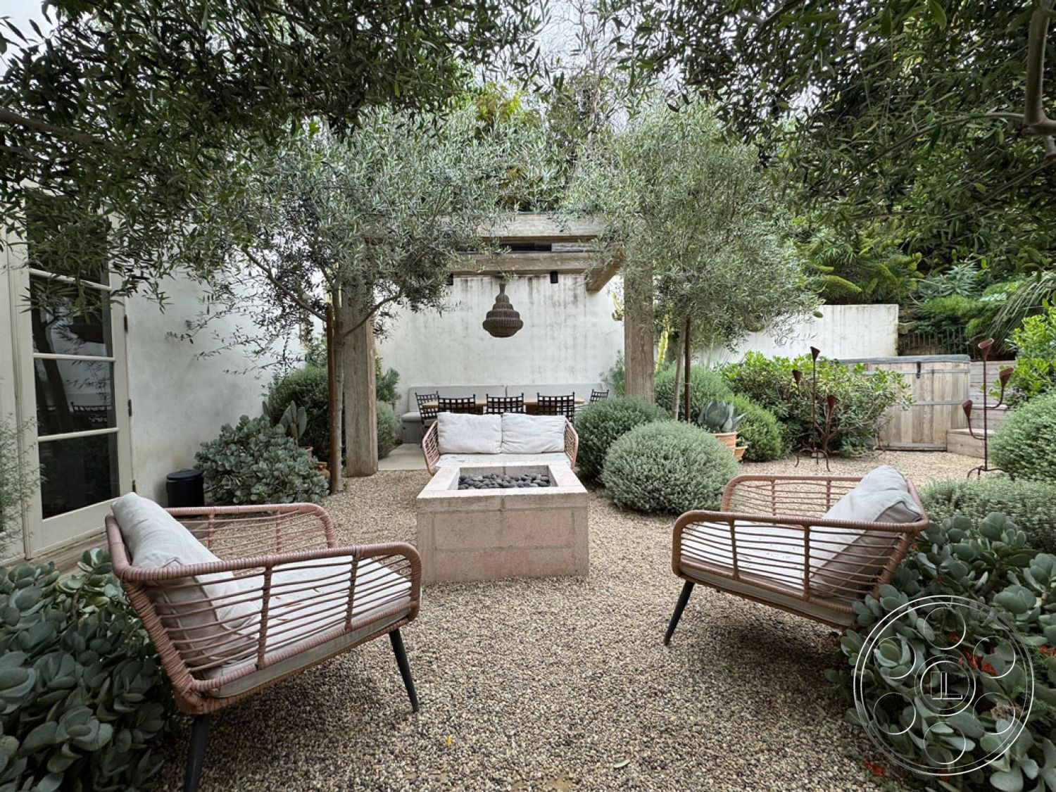 Spanish 165 - exterior, olive trees, modern outdoor space, gravel patio flooring, minimalist garden design, urban garden setting, wooden outdoor dining table, rectangular fire pit, woven rattan patio chairs, concrete planter boxes, green shrub landscaping, natural stone wall backdrop, pergola with hanging light fixture, floral garden accents, open-air relaxation area