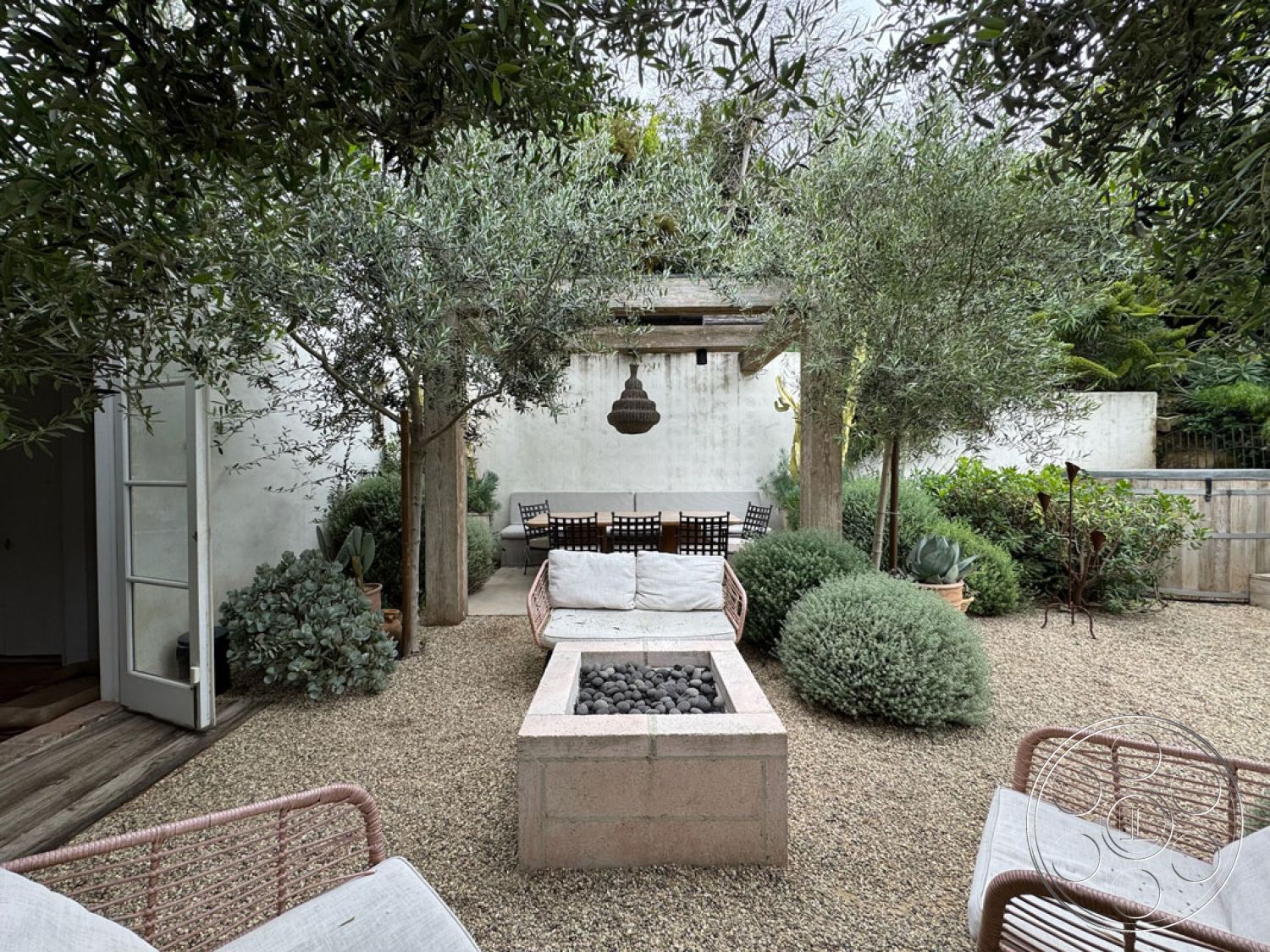 Spanish 165 - exterior, hanging light fixture, minimalist landscaping, gravel patio flooring, private outdoor retreat, concrete fire pit, lush greenery surrounding, wood patio dining table, woven outdoor seating, olive trees in landscape, succulent garden accents, concrete garden walls, modern outdoor design, rustic wood beams, stone garden planters