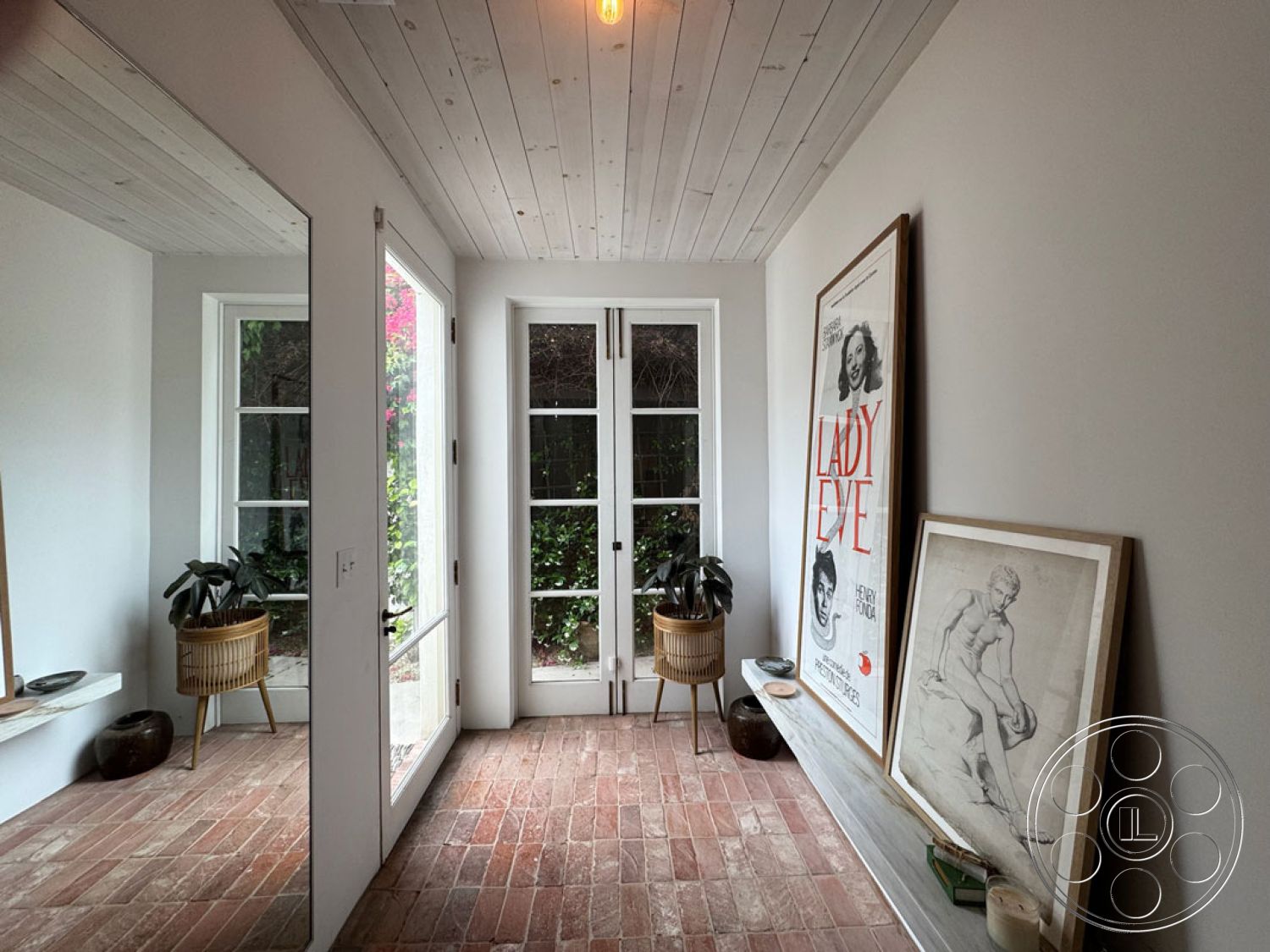 Spanish 165 - neutral color palette, open space layout, glass entry door, natural light from windows, vintage art prints, minimalist interior decor, indoor potted plants, exterior brick flooring, wood plank ceiling detail, framed artwork wall display, textured white painted walls, contemporary entryway design, modern home entrance, woven planter basket, direct garden view