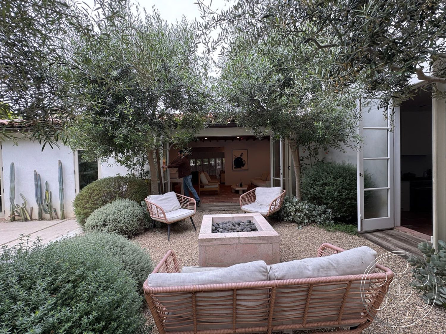 Spanish 165 - open-concept living space, cozy outdoor nook, natural stone fire pit, interior garden patio with gravel flooring, white stucco exterior walls, wooden beam ceiling structure, woven rattan lounge chairs with cushions, olive trees providing shade, decorative cacti wall art, wood sliding doors opening to living area, low maintenance drought-resistant landscaping, brick walkway leading to garden area, modern minimalist outdoor seating, planted shrubs for privacy, suburban backyard retreat