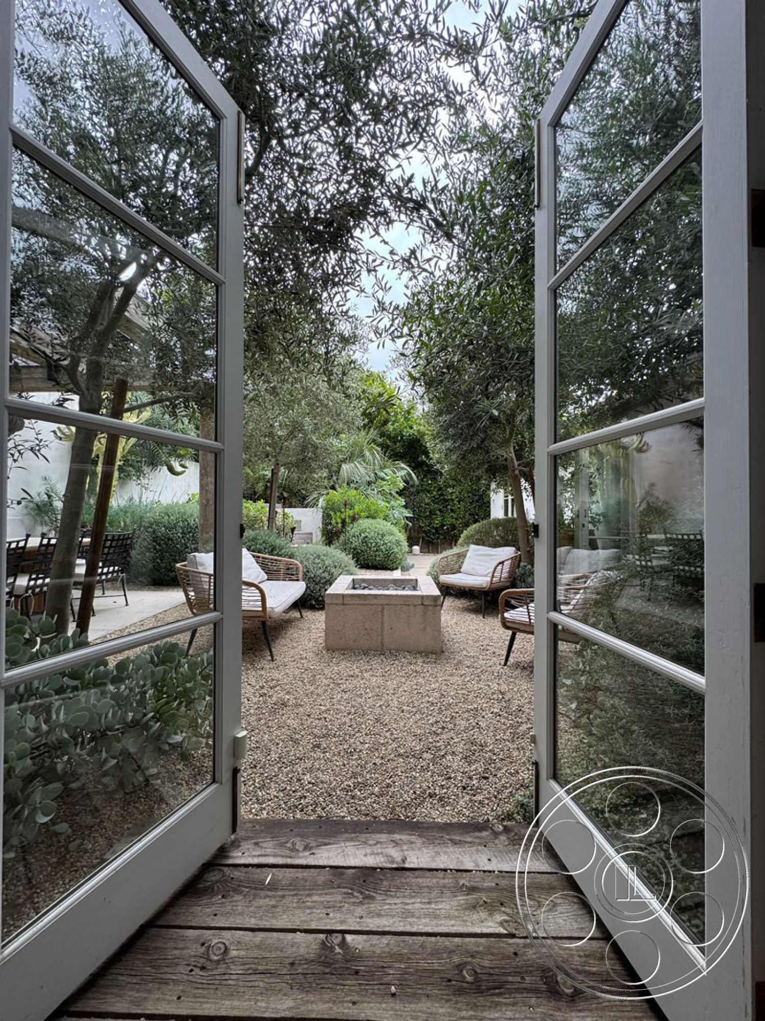 Spanish 165 - outdoor seating area, modern outdoor furniture, stone fire pit, natural stone pavers, landscaped garden setting, wooden door frame, woven rattan chairs, gravel garden floor, double glass doors, olive trees surrounding space, suburban backyard exterior, green shrubbery accents, contemporary fire feature, open-air patio ambiance, rustic wooden deck flooring, textured garden mulch