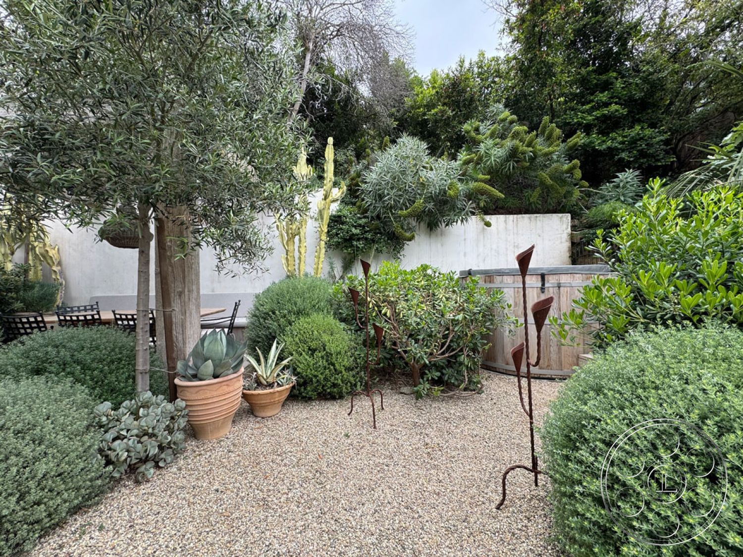 Spanish 165 - privacy wall, outdoor relaxation area, circular planters, exterior garden setting, wooden outdoor dining table, potted succulent plants, natural stone wall backdrop, gravel garden flooring, textured greenery accents, decorative metal garden sculptures, lush shrubbery landscaping, tall cacti accents, organic garden design, mediterranean style plants, wooden wine barrel feature