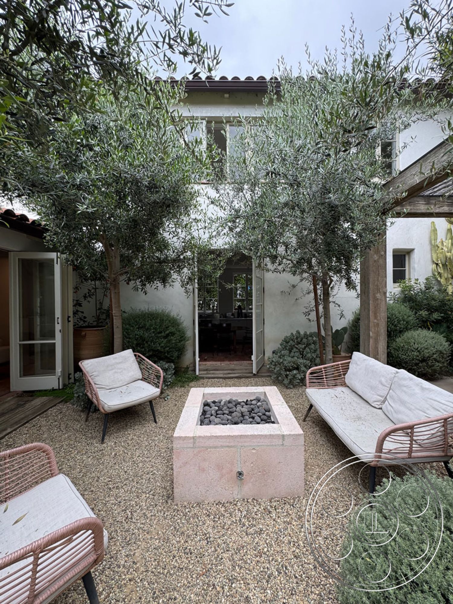 Spanish 165 - gravel patio flooring, modern outdoor seating area, floor-to-ceiling glass doors, wooden pergola structure, exterior courtyard with olive trees, rectangular fire pit with lava rocks, woven patio chairs with light cushions, stucco wall exterior, tropical garden plants, lightweight metal and textile furniture, rustic outdoor aesthetic, urban backyard setting