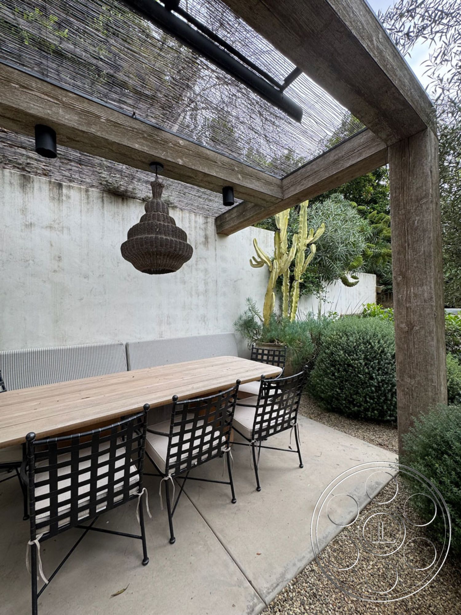 Spanish 165 - textured plaster walls, desert-inspired landscaping, concrete patio flooring, black metal dining chairs, wooden pergola structure, modern outdoor setting, exterior courtyard dining area, reclaimed wood dining table, built-in bench seating with cushions, landscaped garden with native plants, suspended woven light fixture, minimalist outdoor aesthetic, open-air dining space, cactus plant accents, natural stone gravel pathway