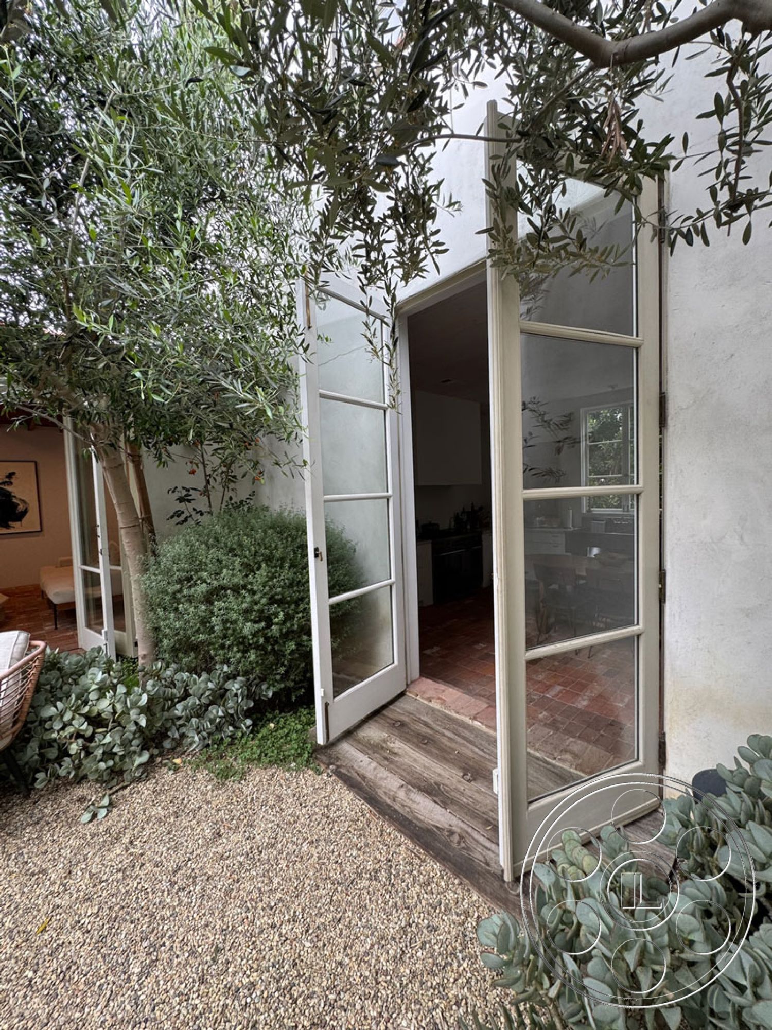 Spanish 165 - lush green landscaping, wooden double doors entrance, gravel pathway exterior, french doors with glass panels, exposed stucco exterior walls, rustic wooden porch flooring, olive trees surrounding entrance, interior view through doors, brick flooring visible inside, ambient natural light from garden, outdoor greenery framing entrance