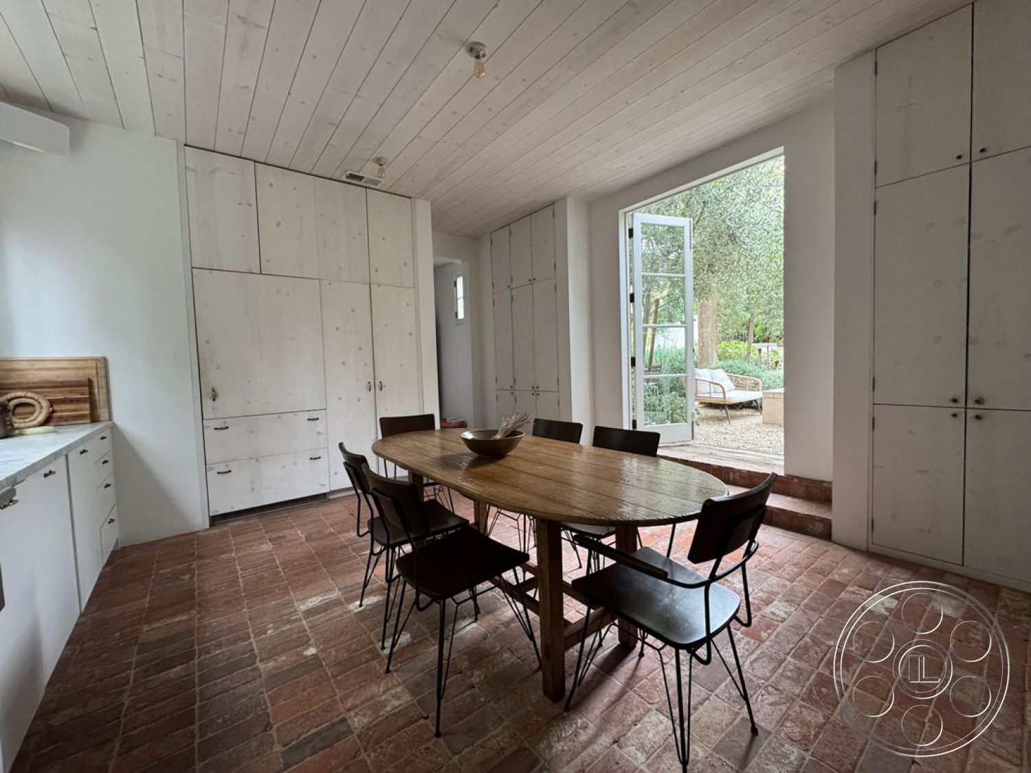 Spanish 165 - wood dining table, earthy color palette, natural wood finishes, arched doorways, textured plaster walls, outdoor garden view, metal dining chairs, serene outdoor setting, interior brick flooring, white painted shiplap ceiling, built-in cabinetry with wooden doors, natural light from open doors, rustic interior design, open floor plan kitchen and dining area, spacious dining room, modern country kitchen, contemporary minimalist style, plank-style ceiling, traditional brick paver flooring