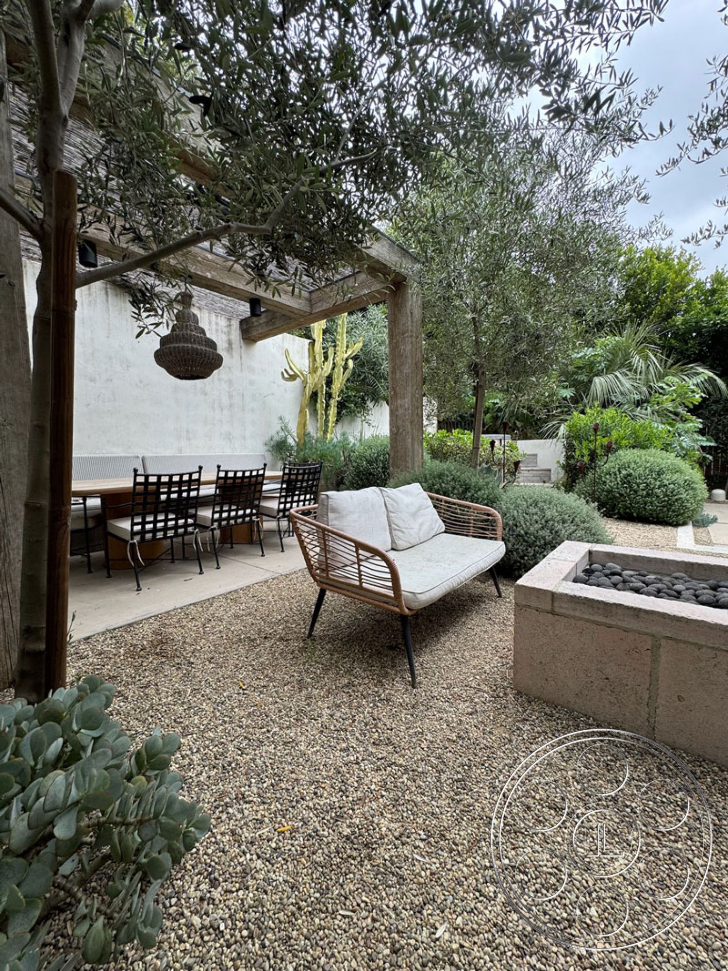 Spanish 165 - neutral color palette, exterior, gravel patio flooring, open outdoor seating area, modern outdoor design, wood pergola with hanging lamp, metal dining table with black chairs, rattan outdoor sofa with cushions, succulent plant landscaping, stone planter box with smooth pebbles, lush desert plants, curved garden paths, natural light filtering through trees, intimate garden setting