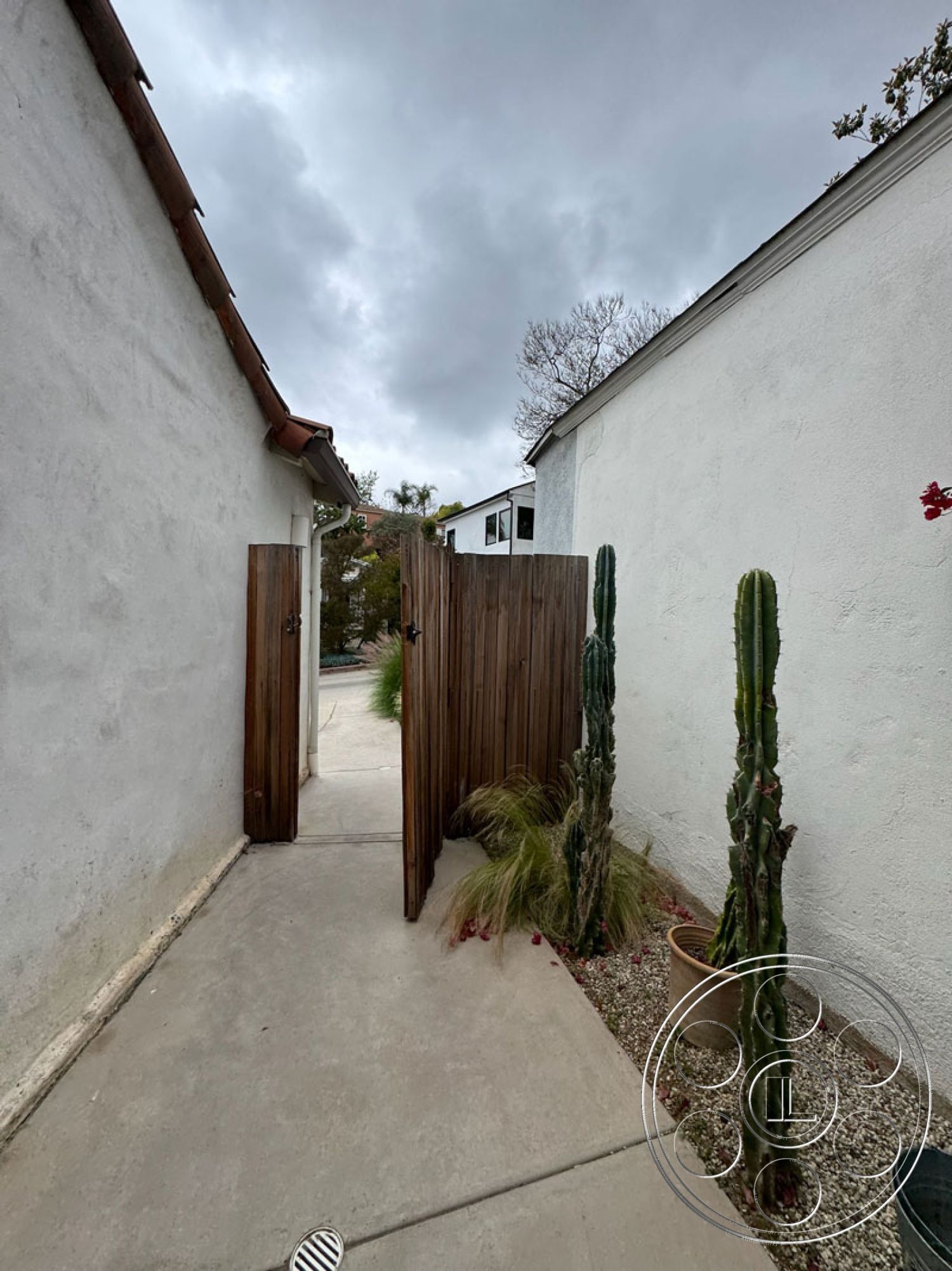 Spanish 165 - exterior, urban setting, concrete walkway, desert landscape, tiled roof, narrow pathway, minimalist landscaping, gravel ground cover, wood fence, stucco wall finish, cloudy sky backdrop, wooden gate entrance, potted cacti, modern residential design, natural plant accents