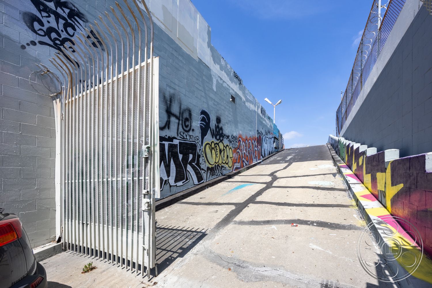 Rooftop 51 - exposed concrete wall, urban setting, exterior concrete driveway, exterior concrete ramp, graffiti wall art detail, metal security gate, sloped street shape, industrial alleyway, surrounding graffiti-covered walls, partial sunlight illumination, asphalt surface detail, concrete curb with colorful mural, high fence perimeter, street lamp features, metal gate design