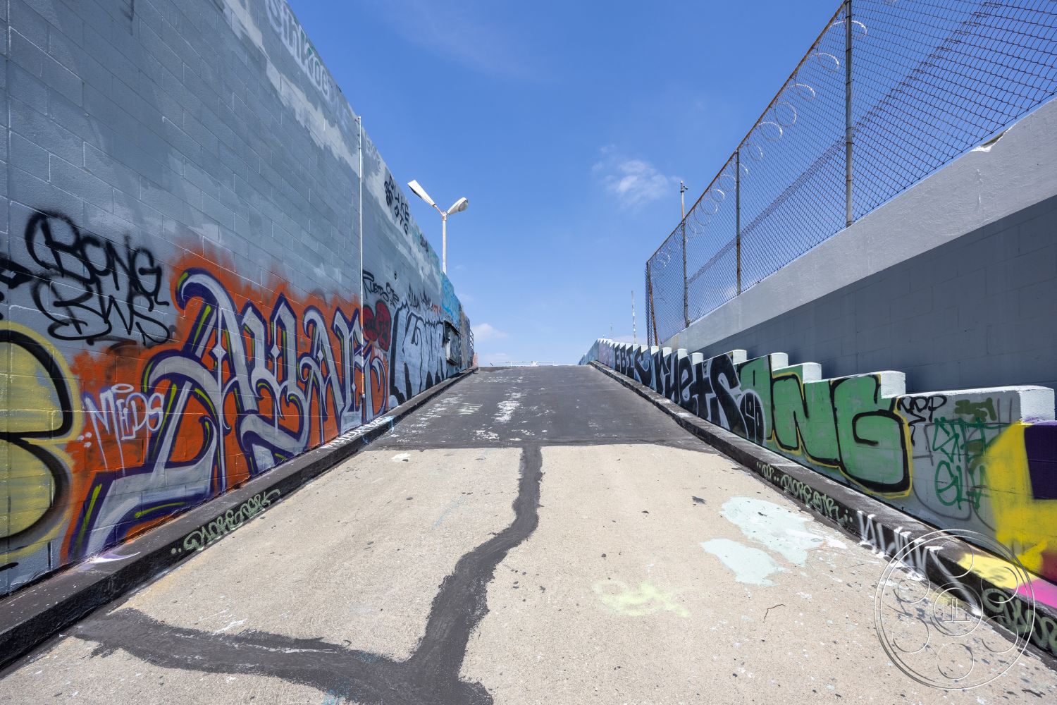 Rooftop 51 - skyline view, textured concrete surface, modern urban architecture, bright blue sky backdrop, surrounding cityscape, exterior concrete ramp, graffiti-covered concrete walls, urban artwork features, asphalt ramp surface, chain link fence detail, industrial-style urban setting, street art elements, sloped access path, exposed concrete block construction