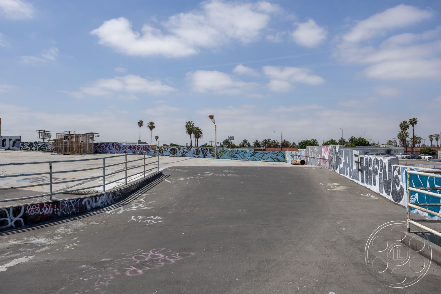 Rooftop 51 - public space, outdoor recreational space, minimal landscaping, metal safety railings, graffiti-covered walls, exterior skate park surface, asphalt skateboarding area, concrete boundary wall, open urban setting, palm tree-lined view, urban art installation, flat surface for skating, contemporary skate park design, bleached sky backdrop, industrial urban landscape