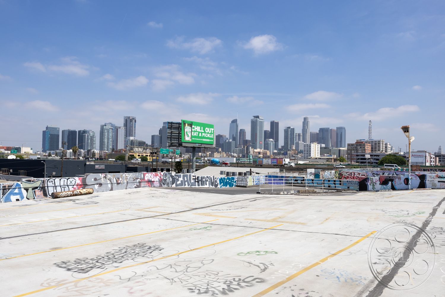 Rooftop 51 - cityscape background, graffiti-covered walls, concrete barriers, exterior rooftop view, concrete rooftop surface, urban city skyline view, industrial warehouse setting, flat rooftop layout, mixed-use urban environment, downtown commercial buildings, metal railings along the rooftop, billboard signage, vehicular parking spaces, open-air rooftop space, concrete surface with spray paint, industrial urban aesthetic
