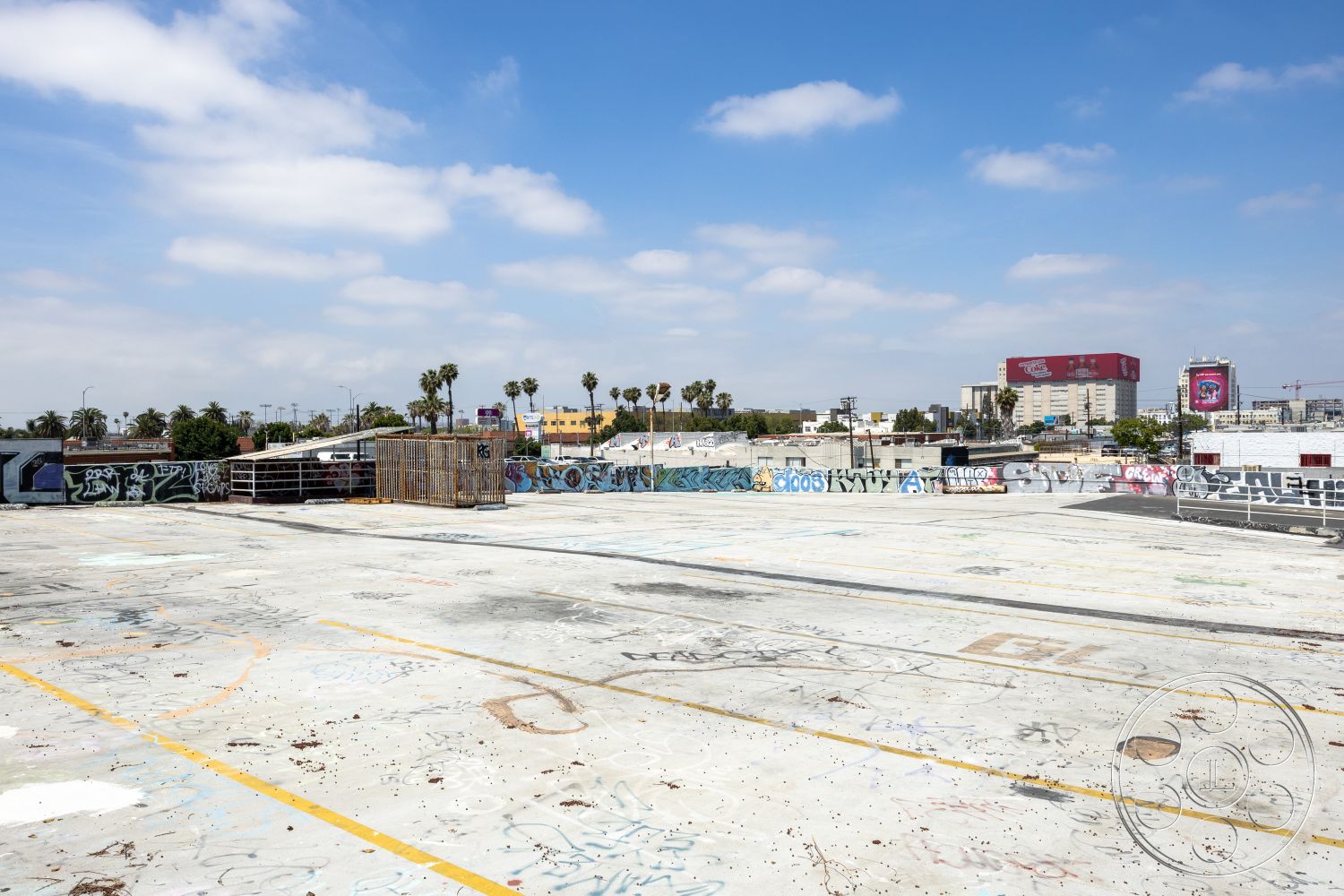 Rooftop 51 - open-air space, city skyline view, wire fencing, distressed concrete texture, flat rooftop design, urban rooftop setting, graffiti-covered walls, urban art installation, concrete rooftop surface, exterior urban landscape, industrial rooftop aesthetic, paved surface with markings, palm trees in vicinity, weathered urban environment, modern cityscape backdrop