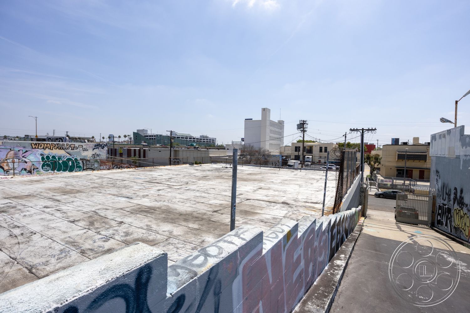 Rooftop 51 - urban skyline view, sunny outdoor environment, asphalt road surface, exposed concrete parking lot surface, commercial building backdrop, industrial urban setting, graffiti wall mural, flat rooftop space, metal fencing perimeter, concrete block boundary wall, utility pole landscaping