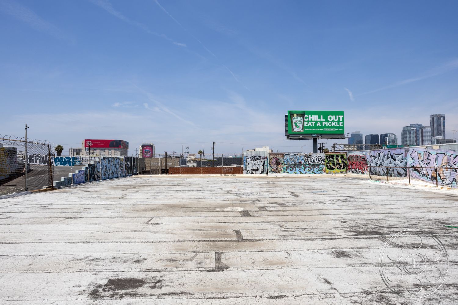Rooftop 51 - city skyline backdrop, exterior concrete rooftop, graffiti-covered walls, billboard signage, urban rooftop view, barbed wire fencing, industrial building setting, weathered concrete surface, open urban space, paved rooftop area