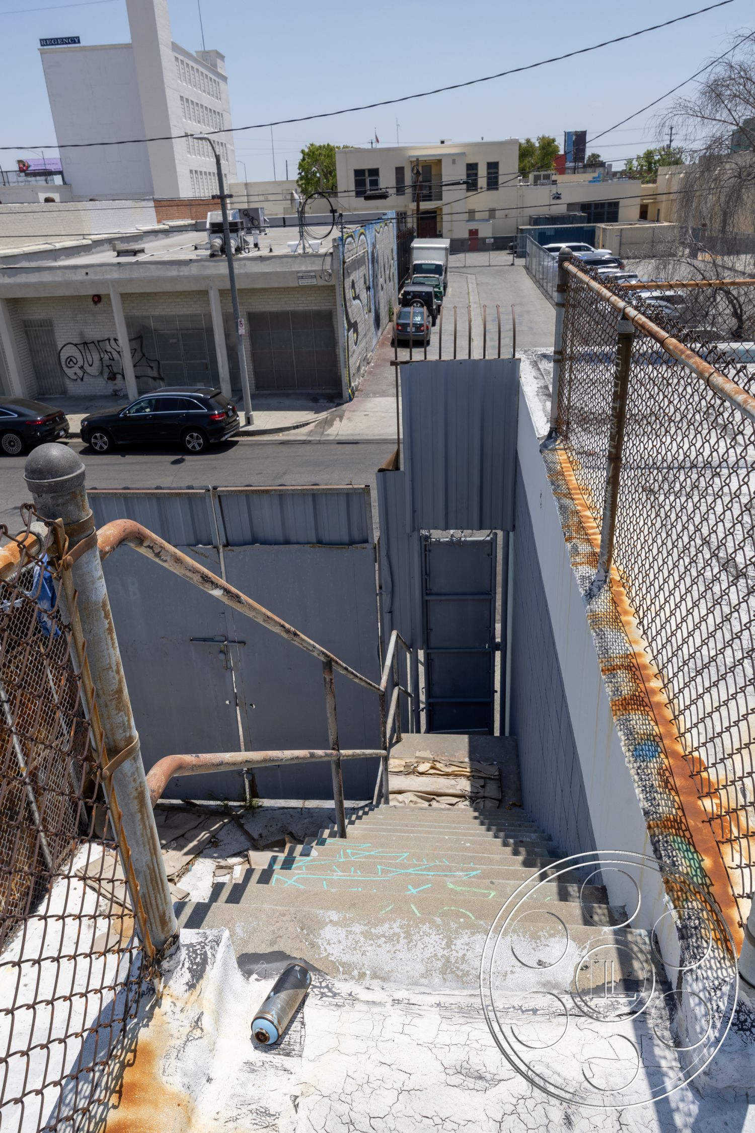 Rooftop 51 - overhead power lines, industrial environment, metal gate entrance, urban parking lot, gray painted walls, chain link fence detail, commercial building backdrop, weathered concrete surface, exterior concrete stairs, exposed metal railing, urban alley setting, industrial style staircase, faded graffiti on walls, asphalt street view, rusted piping details, staircase with worn steps, sidewalk adjacent to stairs