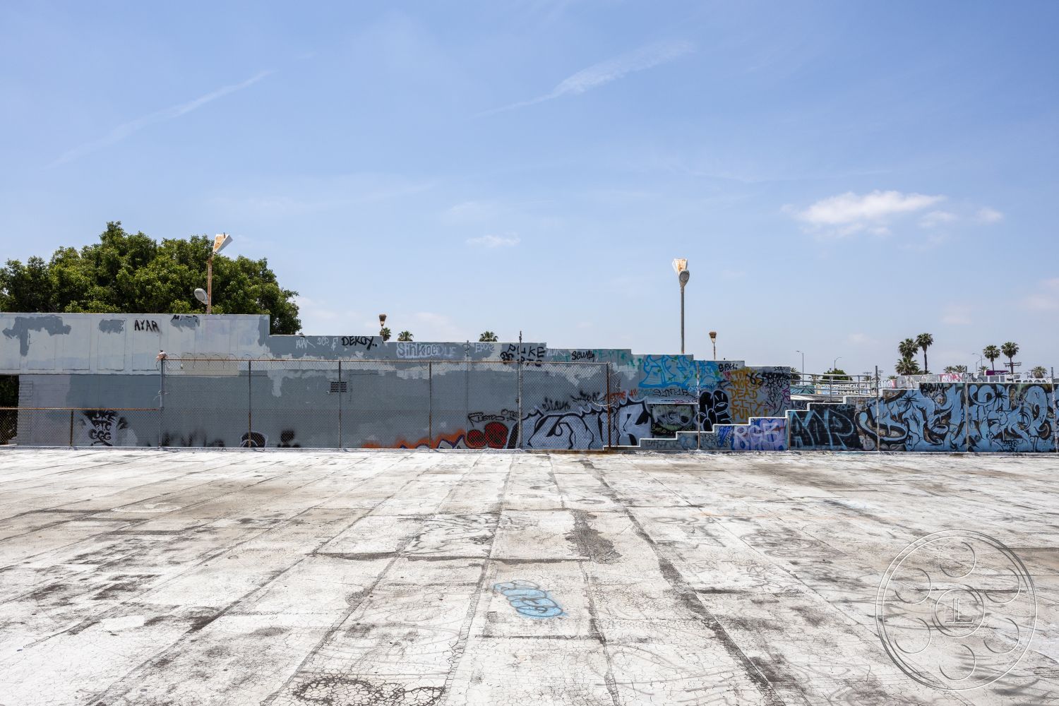 Rooftop 51 - urban landscape, recreational area, chain link fence, industrial setting, flat roof structure, palm tree landscape, asphalt parking area, urban art installation, graffiti wall mural, exterior concrete floor, open space pavement, sunny sky background, commercial building facade, concrete surface wear, metal light posts