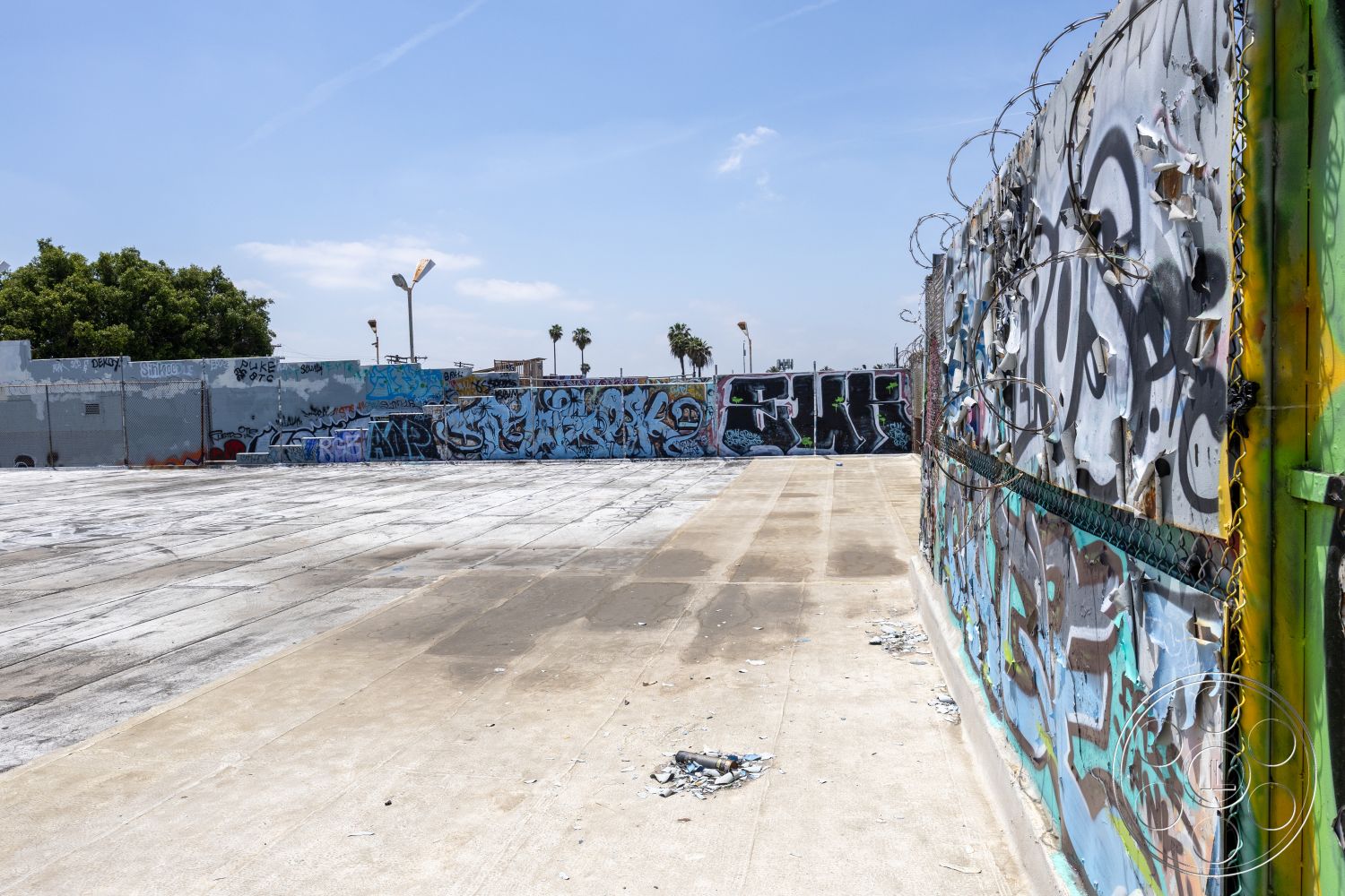 Rooftop 51 - casual gathering area, urban outdoor space, surrounding palm trees, concrete boundary wall, exterior concrete square slab, graffiti-covered chain link fence, paved concrete surface, open industrial lot, exposed barbed wire, scattered debris on pavement, graffiti art details, brightly colored murals, city landscape view, cloudy blue sky backdrop
