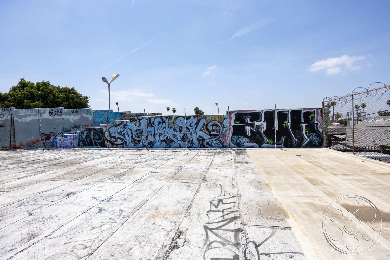 Rooftop 51 - industrial lighting fixtures, cityscape backdrop, surrounding palm trees, exposed aggregate concrete, concrete parking lot surface, graffiti-covered concrete walls, barbed wire fencing, textured concrete finish, urban street art, open outdoor setting, pavement with graffiti marks, suburban industrial environment, exterior urban space