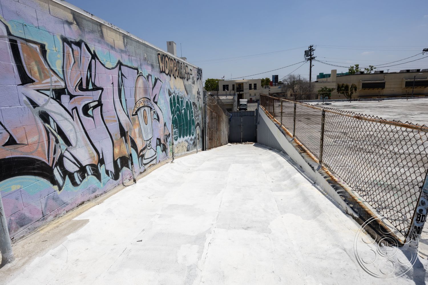 Rooftop 51 - urban landscape, urban environment, paved pathway, textured wall treatment, chain link fence, metal gate entrance, exterior concrete surface, exterior graffiti wall art, industrial alleyway setting, flat roof area, surrounding buildings view, light-colored concrete slope, asphalt backing